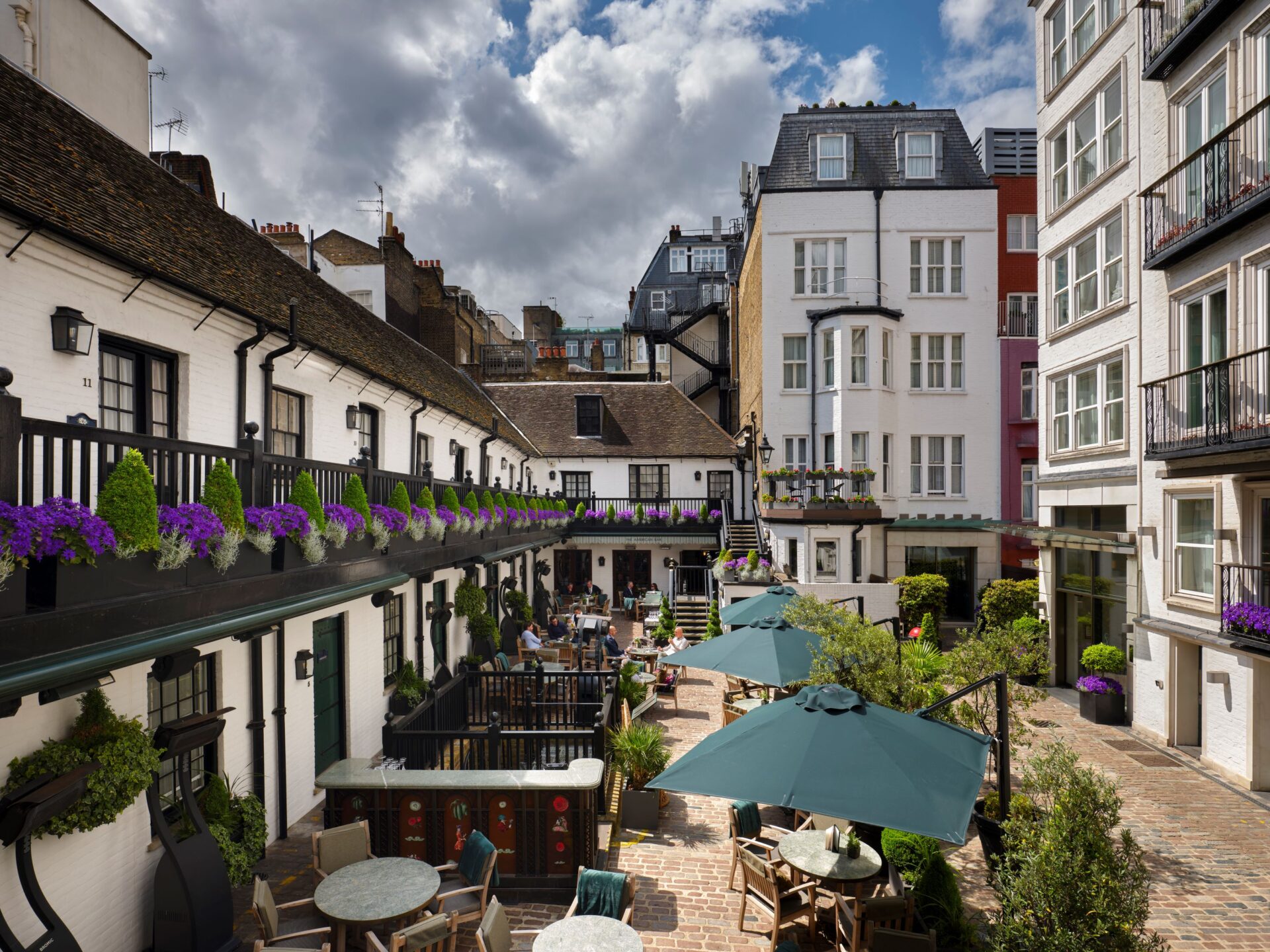 A charming courtyard with outdoor tables, green umbrellas, and potted plants. The area is enclosed by white buildings, some with flower boxes, and features people dining under a partly cloudy sky.