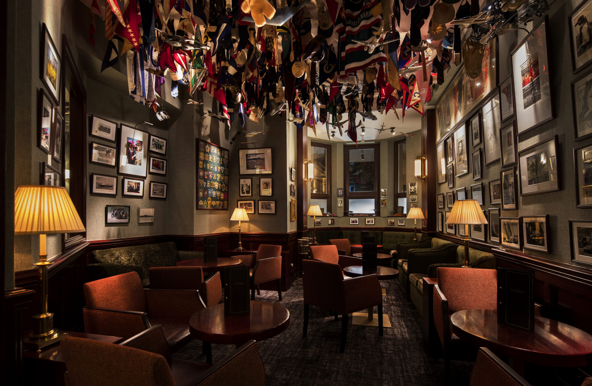 A warmly lit lounge with armchairs and round tables, walls covered in framed photos, and a ceiling decorated with hanging flags and pennants. Table lamps create a cozy, inviting atmosphere.