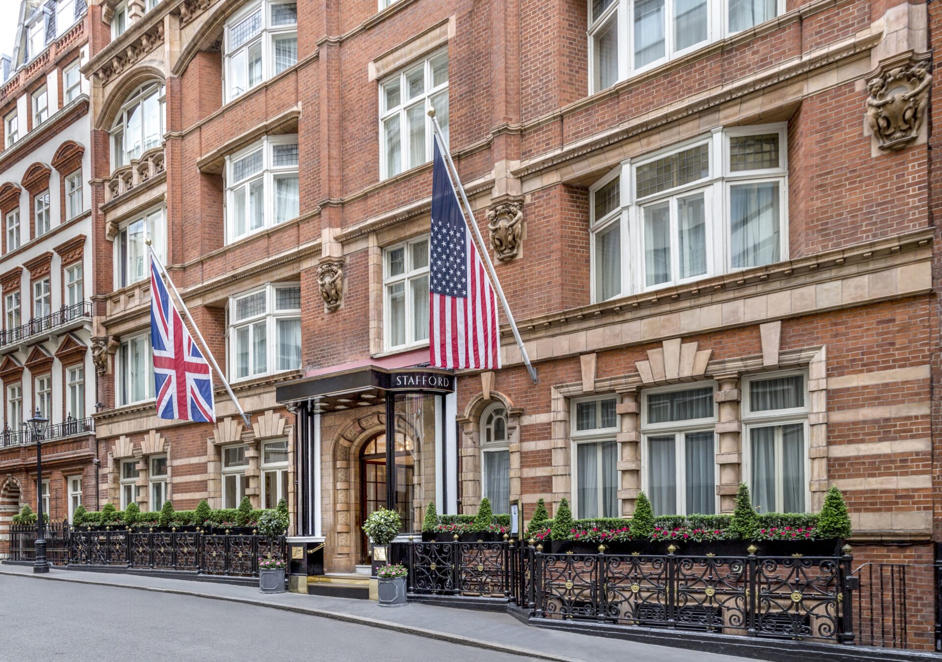 The exterior of a red-brick building with large white-trimmed windows, featuring the British and American flags at the entrance and a sign reading STAFFORD above the door, welcomes guests with black railings and exclusive room offers.