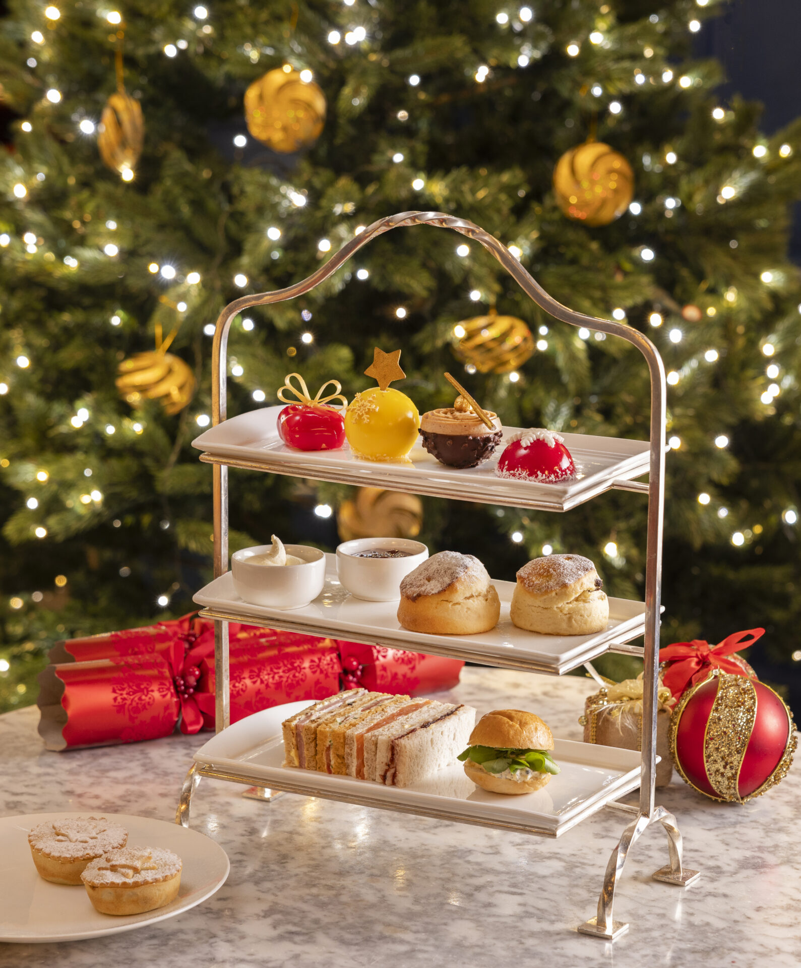 Enjoy a festive afternoon tea set with assorted pastries, scones, and sandwiches elegantly arranged on a three-tiered stand, all set against a backdrop of Christmas tree and holiday decorations.