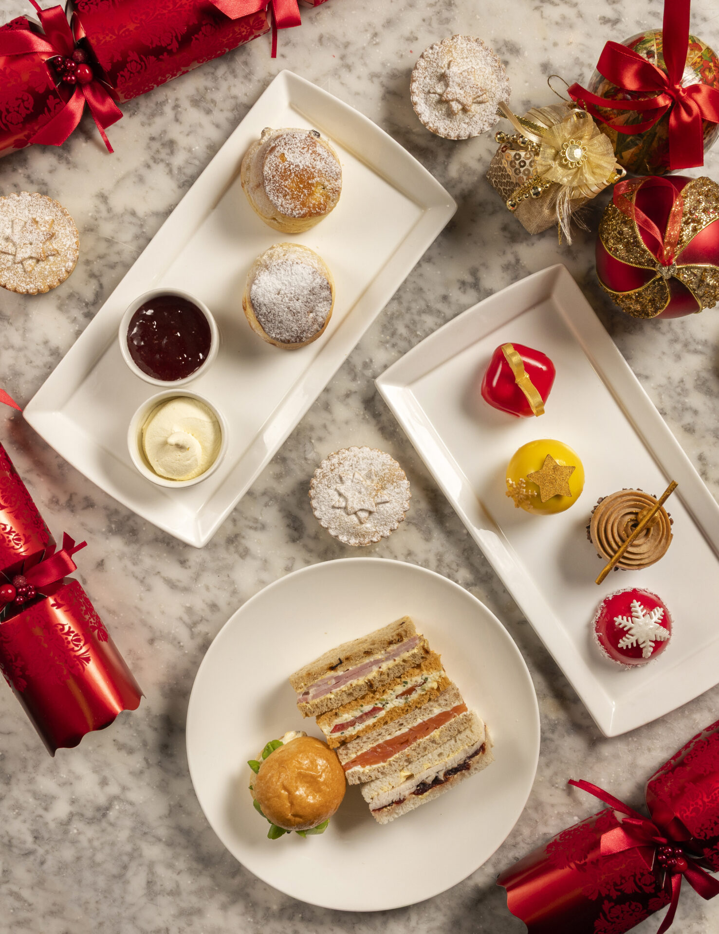 A festive afternoon tea spread on a marble table with Christmas crackers, pastries, jam and cream, colorful desserts, a sandwich cake slice, and a savory bun. Gold and red decorations add a holiday touch.