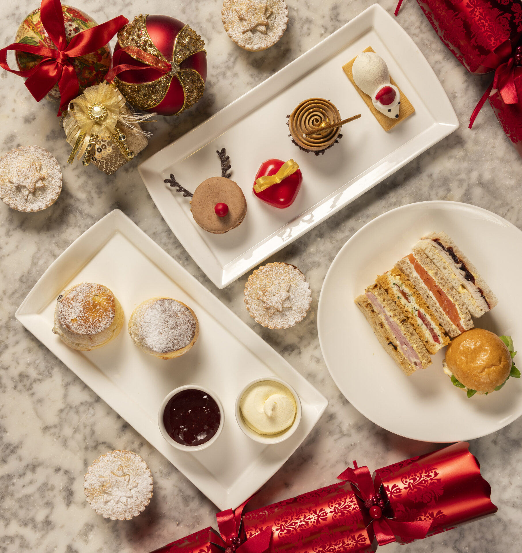 Three plates on a marble table hold assorted festive desserts, pastries, a mini sandwich, and a scone with jam and cream—perfect for afternoon tea. Decorative Christmas ornaments and crackers are arranged around the plates.