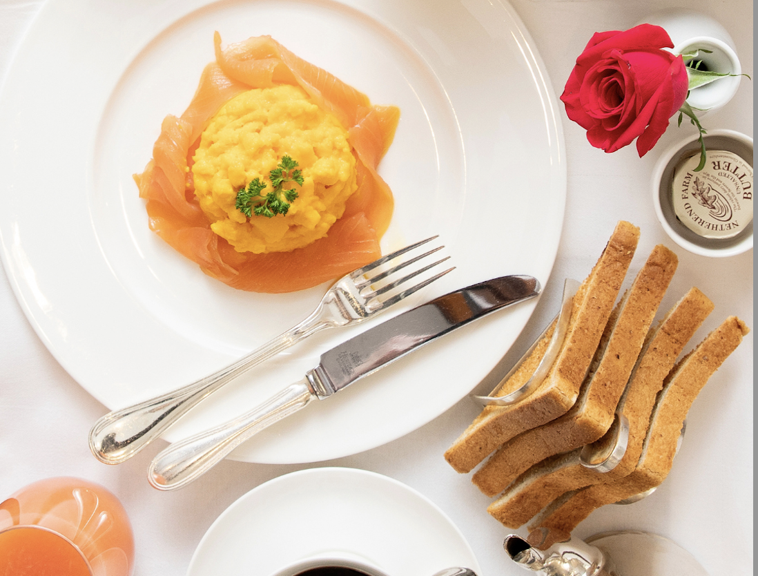 A plate of scrambled eggs on smoked salmon, inspired by The Game Bird menus, is garnished with parsley and served with toast, butter, a cup of coffee, a red rose, and orange juice on a white tablecloth.