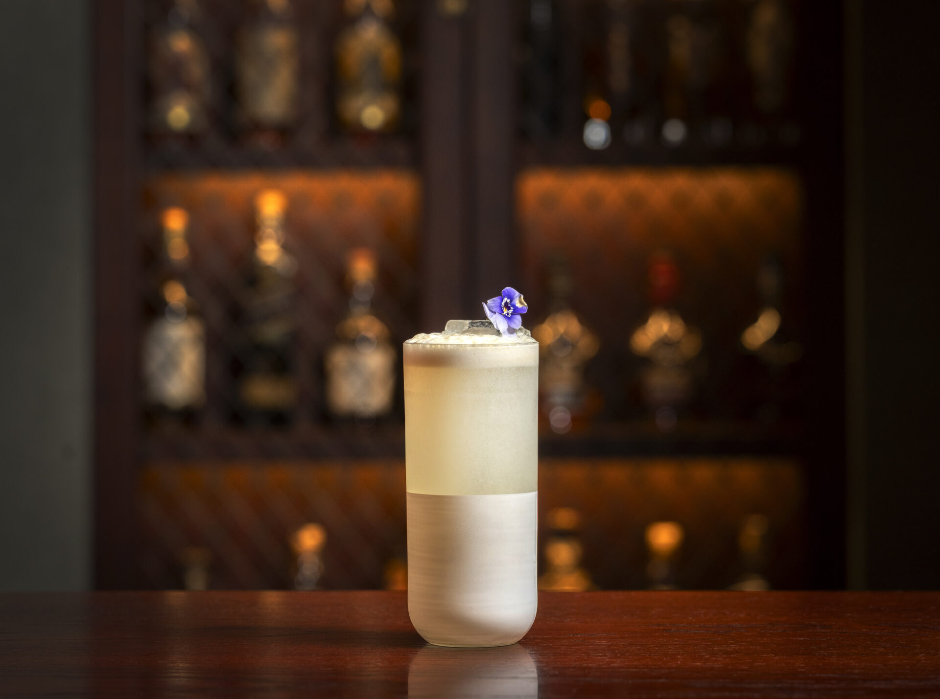 A creamy cocktail garnished with a purple edible flower sits on the wooden bar counter at The American Bar, with a softly lit shelf of liquor bottles blurred in the background.