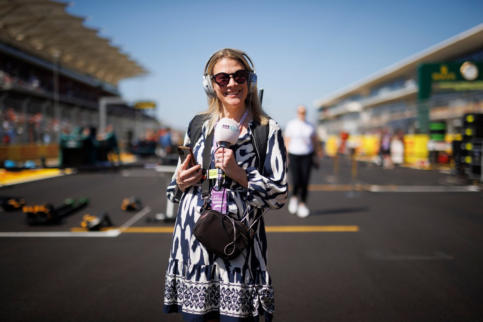A woman wearing sunglasses and headphones holds a microphone and phone while standing on a racetrack, reminiscent of the vibrant atmosphere at The Stafford London. Behind her, blurred people and racing equipment shine under the bright sun and clear blue sky.