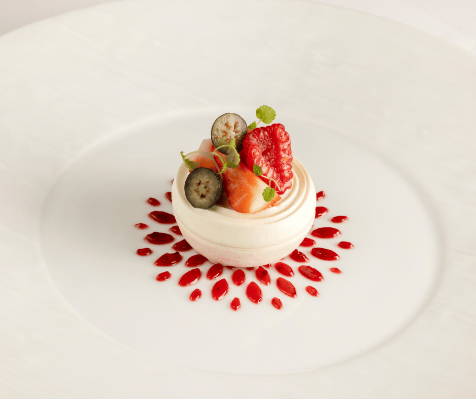 A small meringue dessert topped with fresh berries and garnished with mint leaves, placed on a white plate with a decorative ring of red berry sauce dots around it.