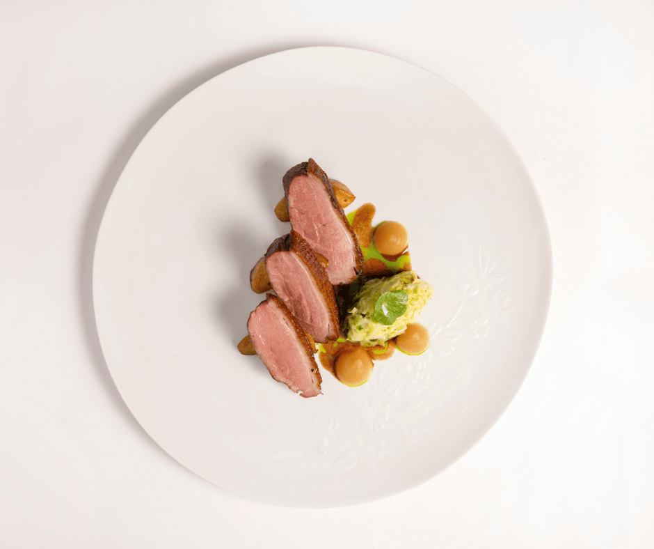 Two slices of seared duck breast with a crispy edge, served with a green-topped scoop of mashed potatoes and dollops of brown sauce, artfully arranged at Michael Caines at The Stafford on a large white plate against a white background.