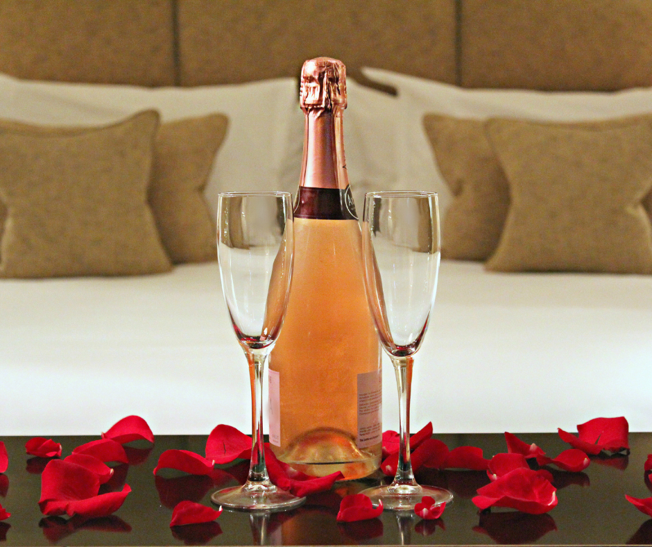 A bottle of rosé champagne and two empty flutes are placed on a table covered with red rose petals in front of a neatly made bed with brown and white pillows.