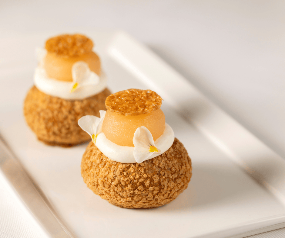 Two elegant pastry puffs topped with cream, pear slices, small crisp discs, and white edible flower petals—an exquisite treat perfect for afternoon tea—arranged on a white rectangular plate against a soft background.