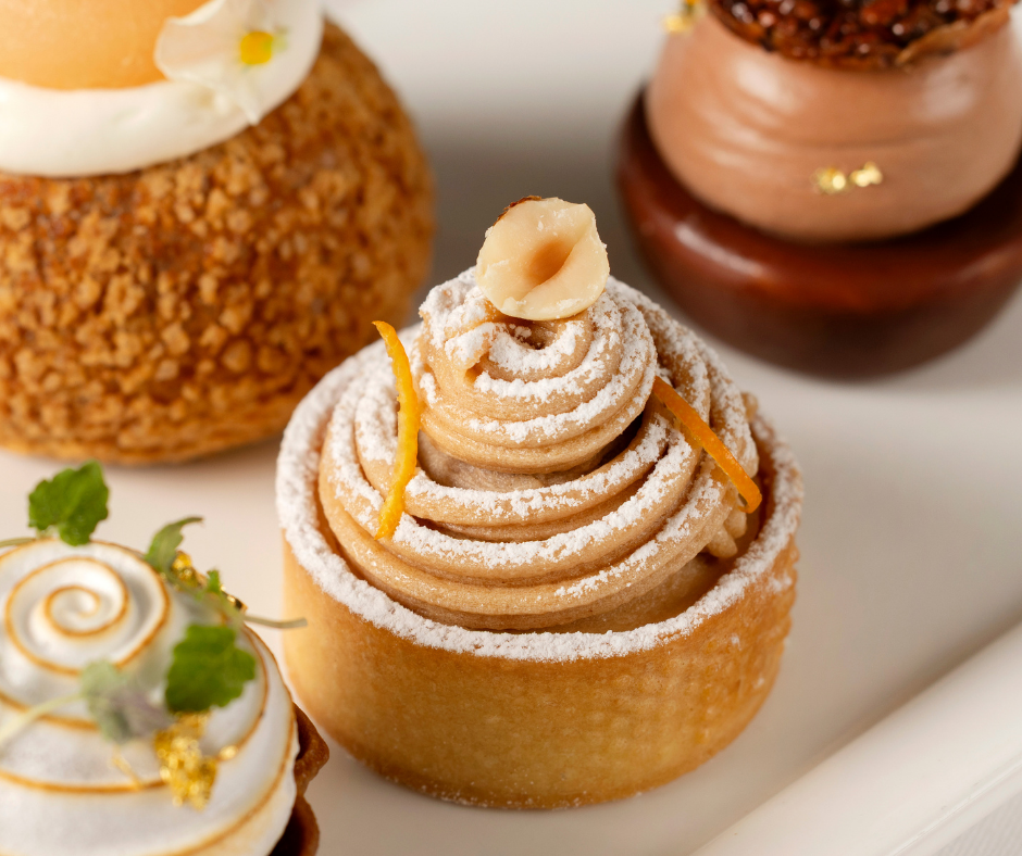 A close-up of assorted elegant pastries perfect for afternoon tea, featuring a tart topped with swirled cream, powdered sugar, and a hazelnut, surrounded by other beautifully decorated desserts on a white plate.
