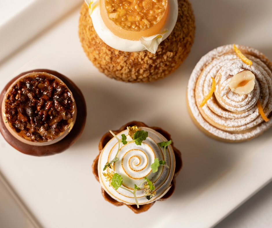 Four assorted gourmet pastries are arranged on a white plate, each decorated with unique toppings such as nuts, cream, citrus zest, and mint leaves—perfect treats for an elegant afternoon tea. The image is taken from above, showcasing their intricate designs.