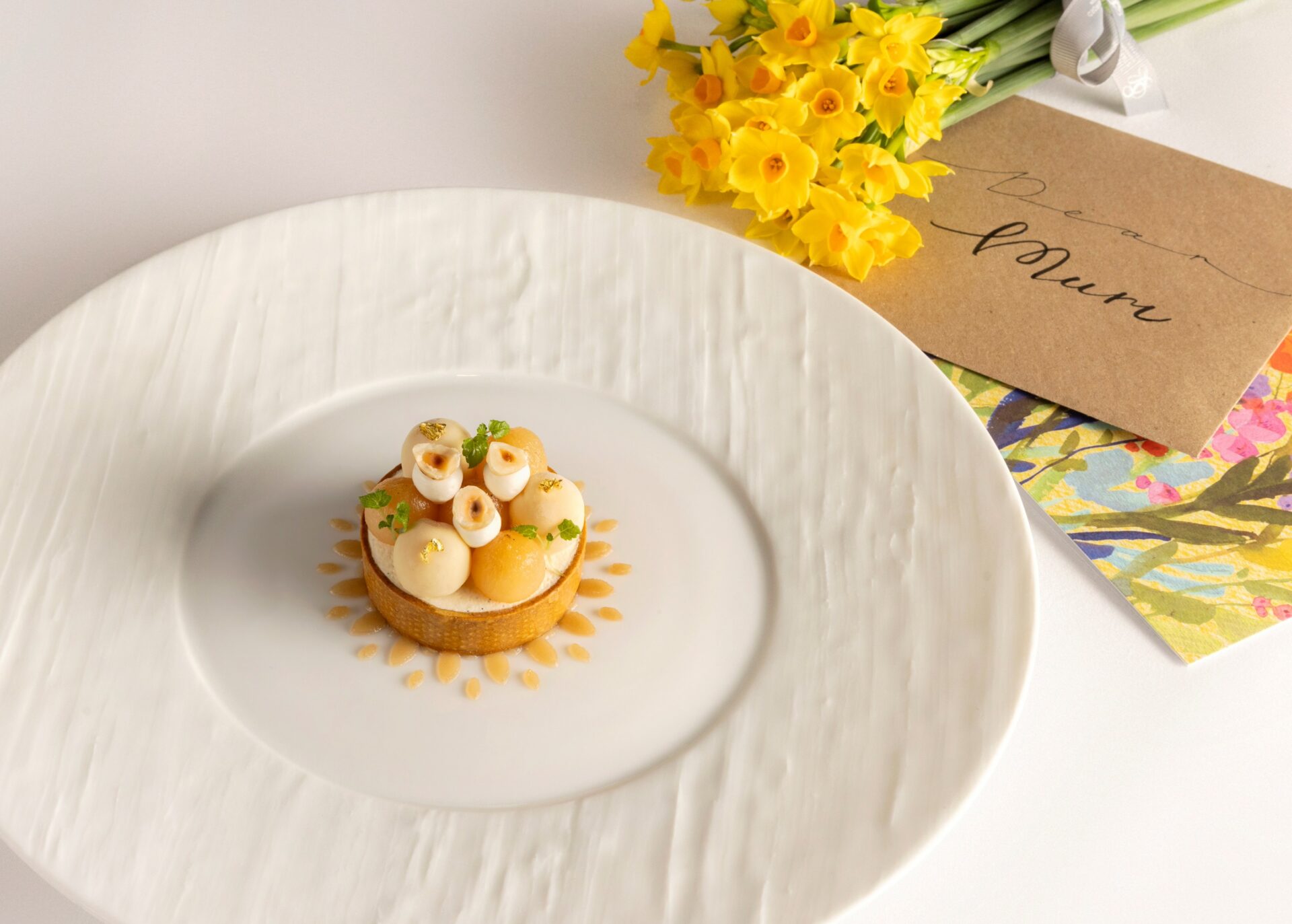A decorative dessert tart on a white plate, next to a bouquet of yellow daffodils and greeting cards, one labeled Dear Mum, on a white table.