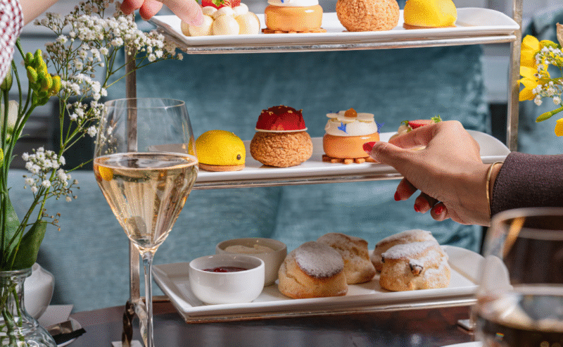 Two people celebrating Mother's Day with an elegant afternoon tea, sharing a tiered tray of colorful pastries, scones, and desserts, alongside glasses of wine, tea, and fresh flowers on a wooden table.