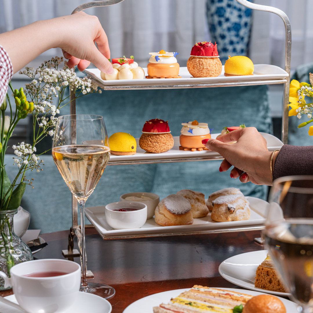 Two people celebrating Mother's Day with an elegant afternoon tea, sharing a tiered tray of colorful pastries, scones, and desserts, alongside glasses of wine, tea, and fresh flowers on a wooden table.