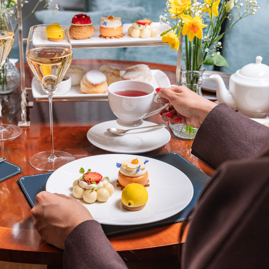 A person enjoys afternoon tea at a table set with elegant pastries on a white plate, a tiered tray of desserts, a glass of wine, a teapot, and a vase of yellow flowers while holding a teacup.