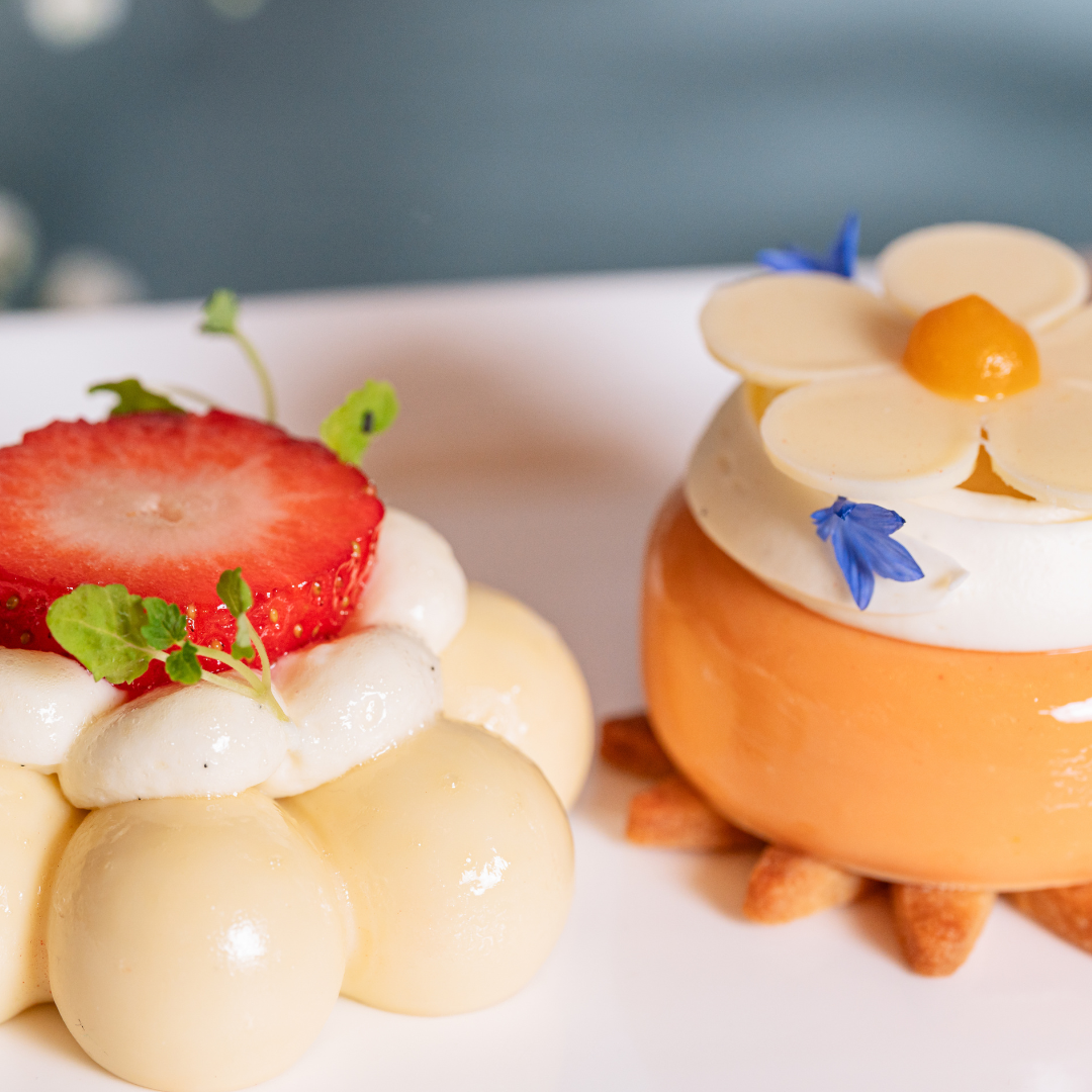 Two elegant Mother’s Day desserts: one topped with a strawberry slice and microgreens, featuring glossy cream dollops; the other is orange, crowned with a white chocolate flower, yellow center, and blue edible petals on a white plate.