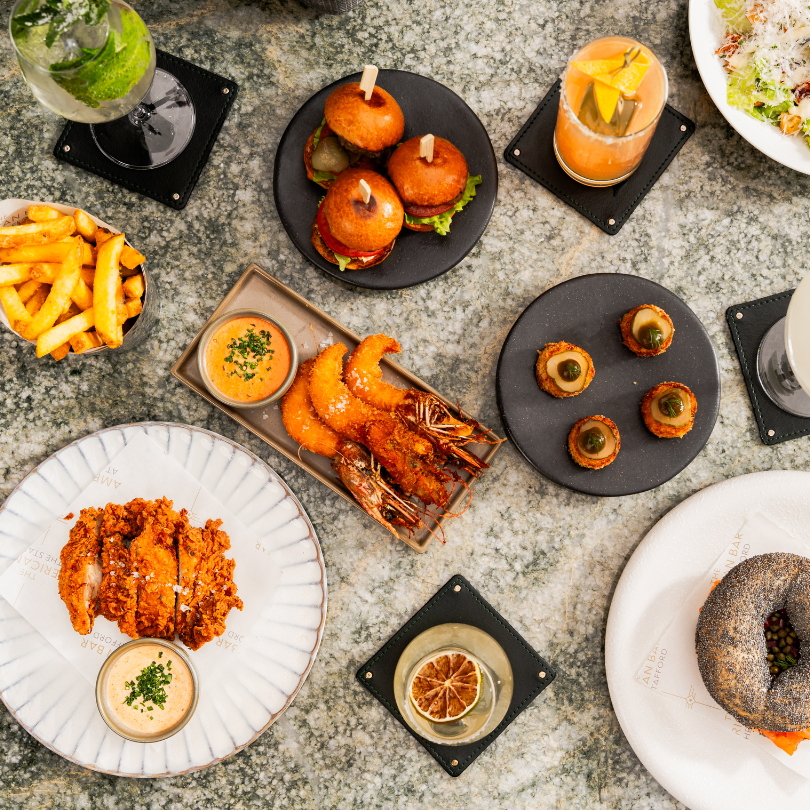 An overhead view of a table at Enemigo Tequila Courtyard showcases burgers, fries, fried shrimp, a breaded cutlet, appetizers, a poppy seed bagel, and assorted drinks with garnishes.