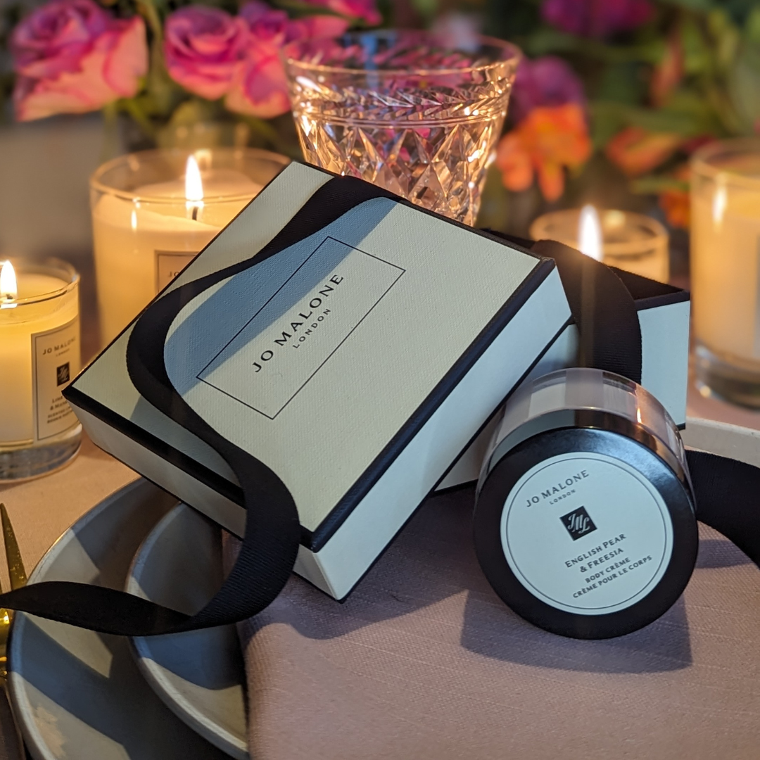 A Jo Malone London gift box with a black ribbon and an English Pear & Freesia body cream jar are displayed on a plate. Lit candles, pink flowers, and hints of wine tasting in the background create an elegant, cozy setting.