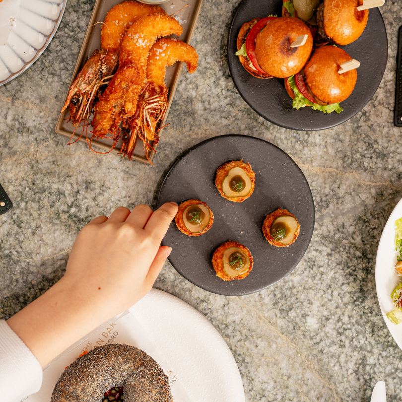A hand reaches for an appetizer on a table in the Enemigo Tequila Courtyard, with shrimp, mini burgers, and bite-sized snacks arranged on plates over a gray marble surface.