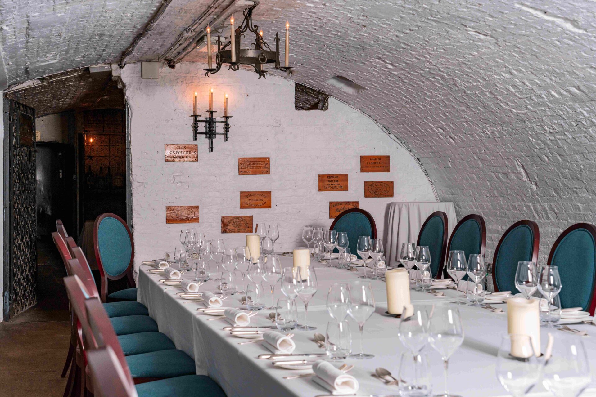 Elegant dining room in a brick-walled cellar at The Stafford London, featuring a long table set with white linens, candles, wine glasses, and teal-upholstered chairs. Chandeliers and wall sconces provide soft lighting.
