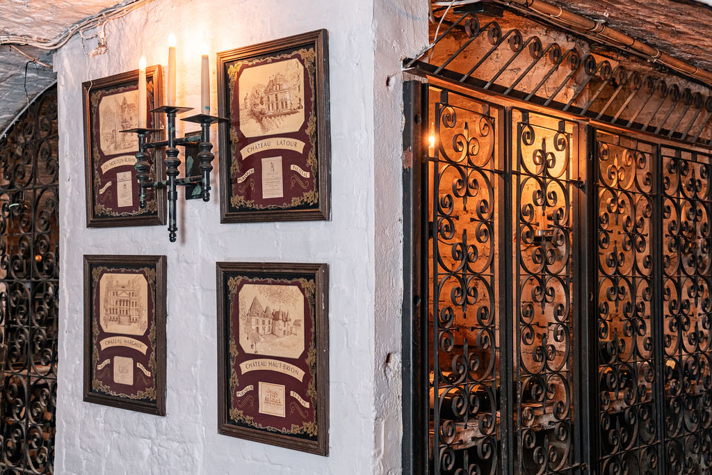 A white brick wall with four framed vintage-style illustrations and a black metal wall sconce with candles sets the mood for wine tasting, next to decorative wrought iron gates in a dimly lit, rustic interior.