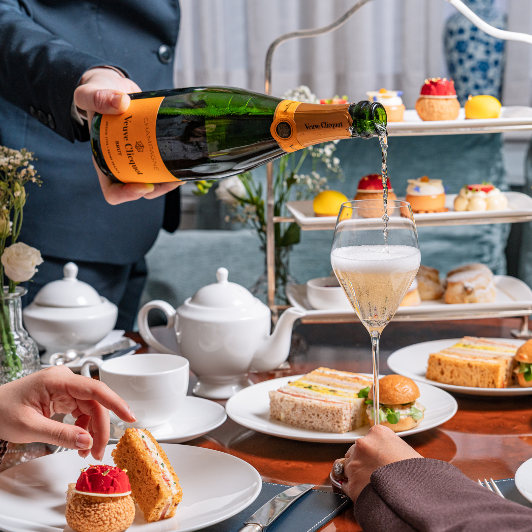 A person pours Veuve Clicquot champagne into a glass at a table set for afternoon tea, surrounded by sandwiches, pastries, teapots, and tiered trays of desserts. Two people are seated, reaching for food and enjoying the elegant afternoon tea spread.
