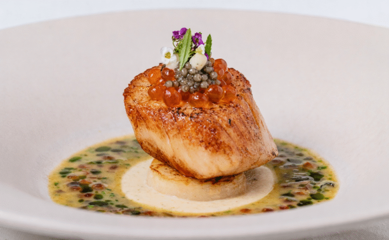 A seared scallop sits atop a round base, garnished with orange and black caviar, microgreens, and edible flowers reminiscent of the Chelsea Flower Show, all surrounded by a creamy, herb-flecked sauce in a white bowl.