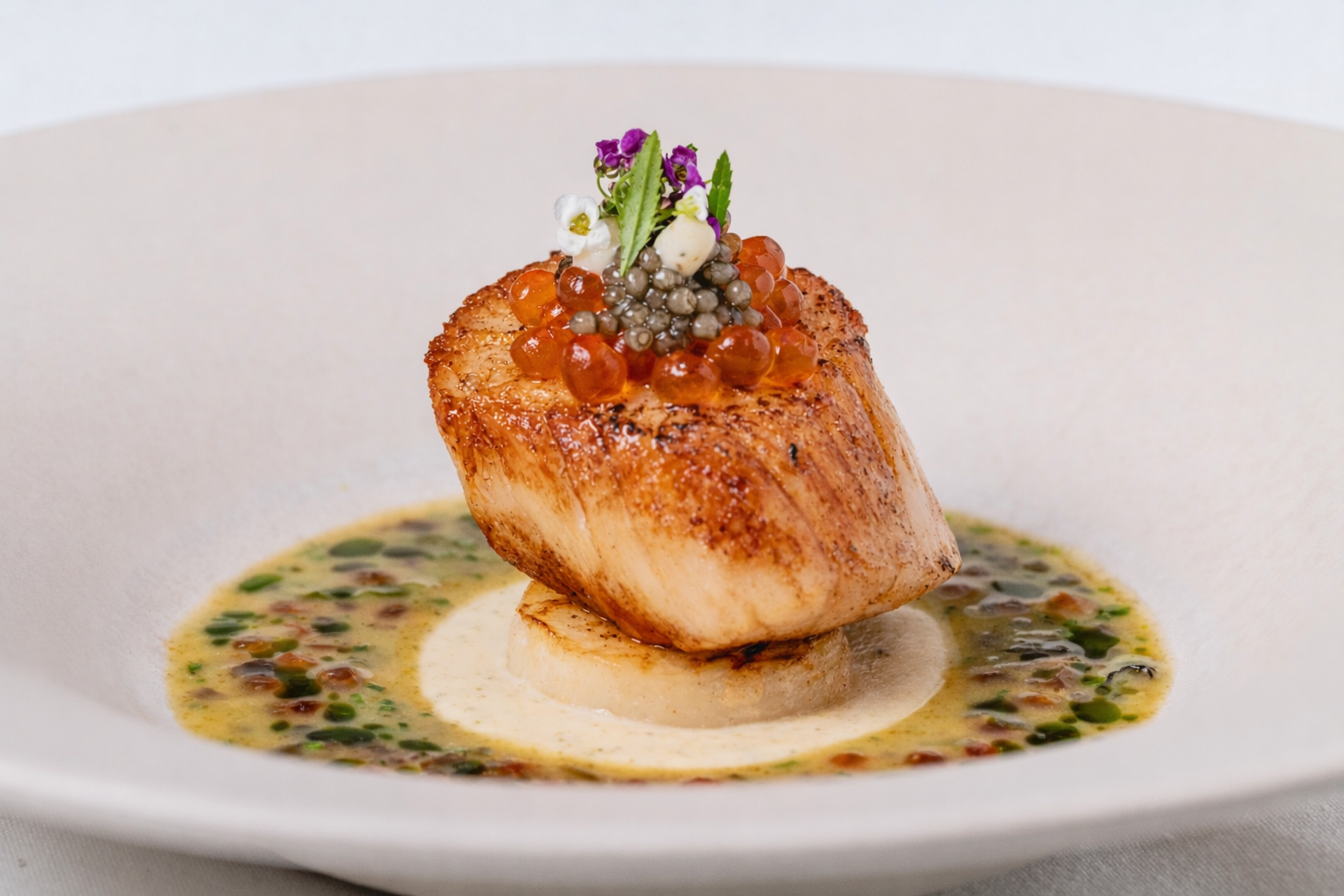 A seared scallop sits atop a round base, garnished with orange and black caviar, microgreens, and edible flowers reminiscent of the Chelsea Flower Show, all surrounded by a creamy, herb-flecked sauce in a white bowl.