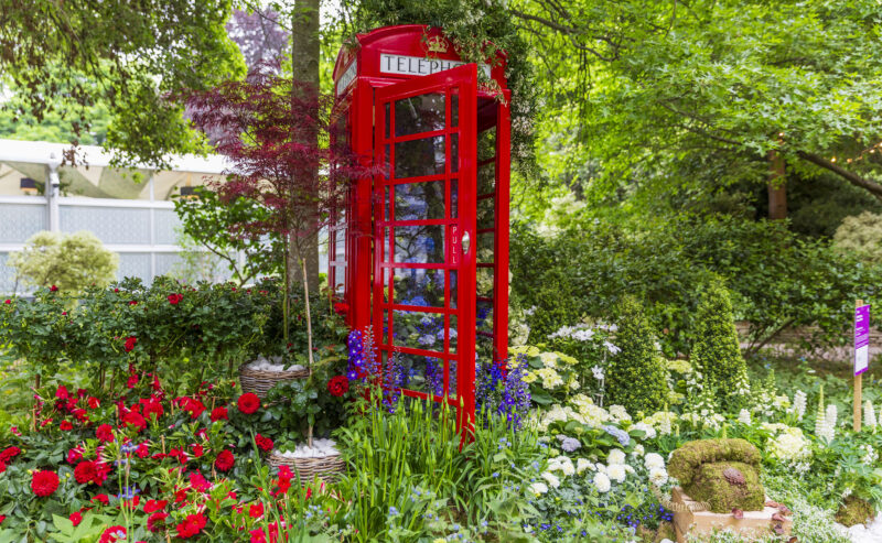 A bright red British telephone booth stands amid a vibrant garden filled with colorful flowers, green shrubs, and trees, creating a whimsical, lush scene perfect for a charming Mother's Day stroll.