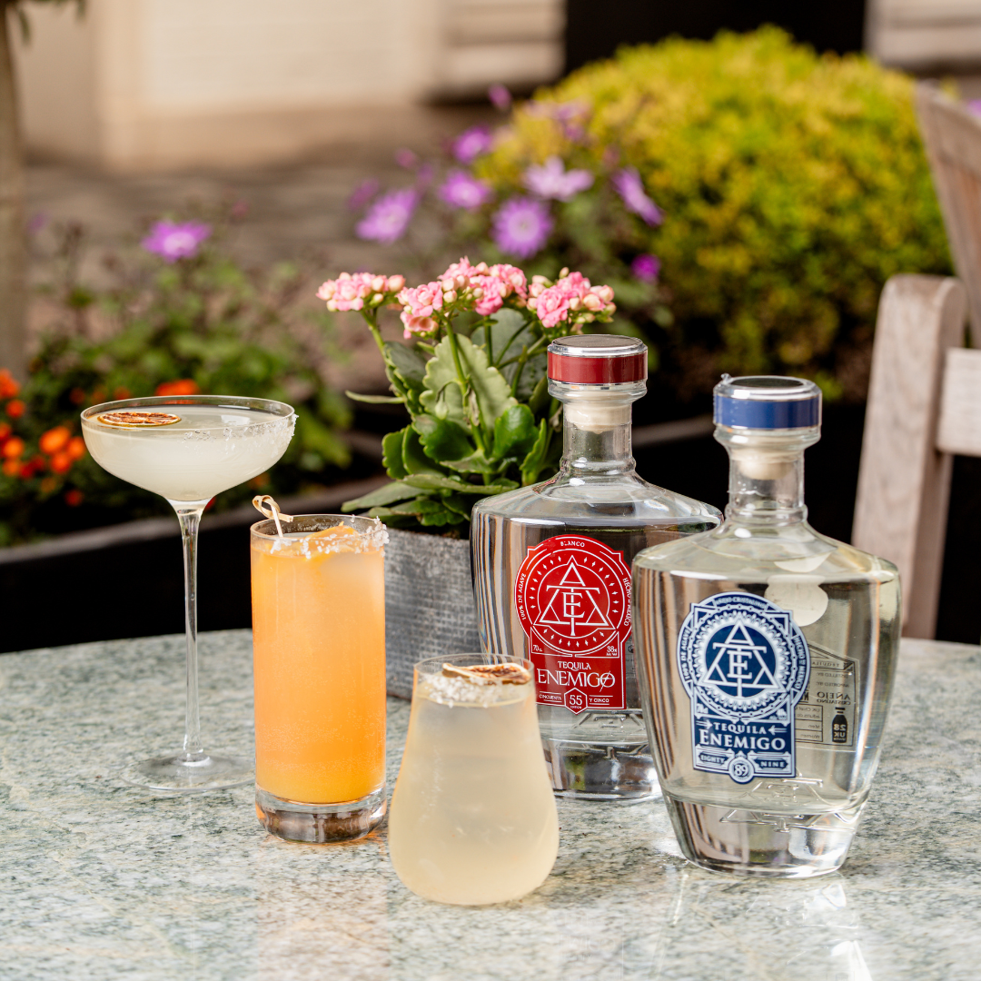 Three tequila cocktails are arranged on a marble table at The American Bar, alongside two Tequila Enemigo bottles and a potted plant. A blurred garden with colorful flowers appears in the background.