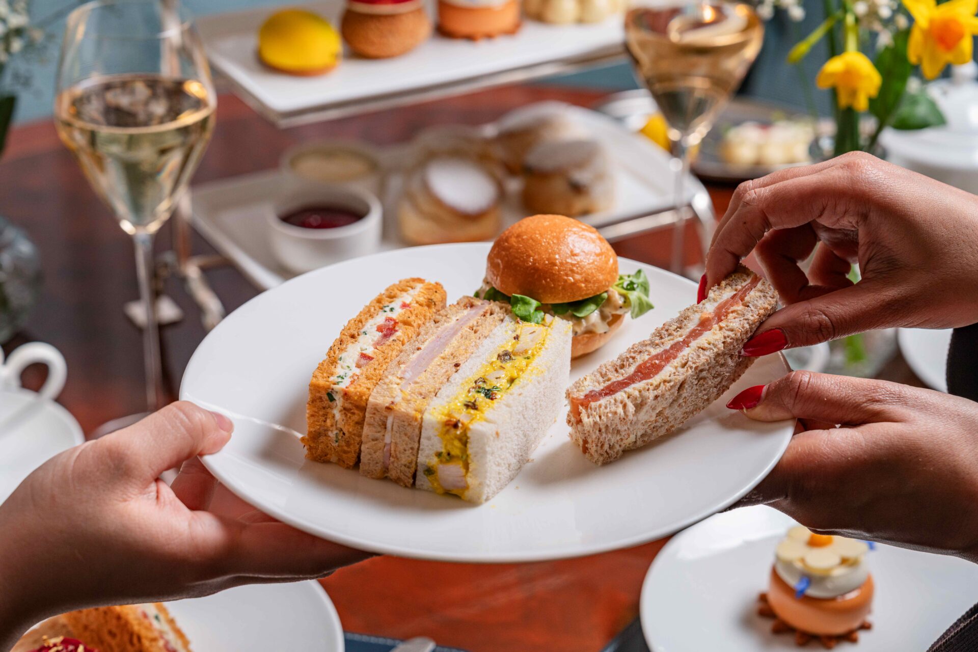 A close-up of two hands holding a white plate with assorted finger sandwiches and a small slider, perfect for afternoon tea, with more tea snacks, desserts, and glasses of white wine in the blurred background.