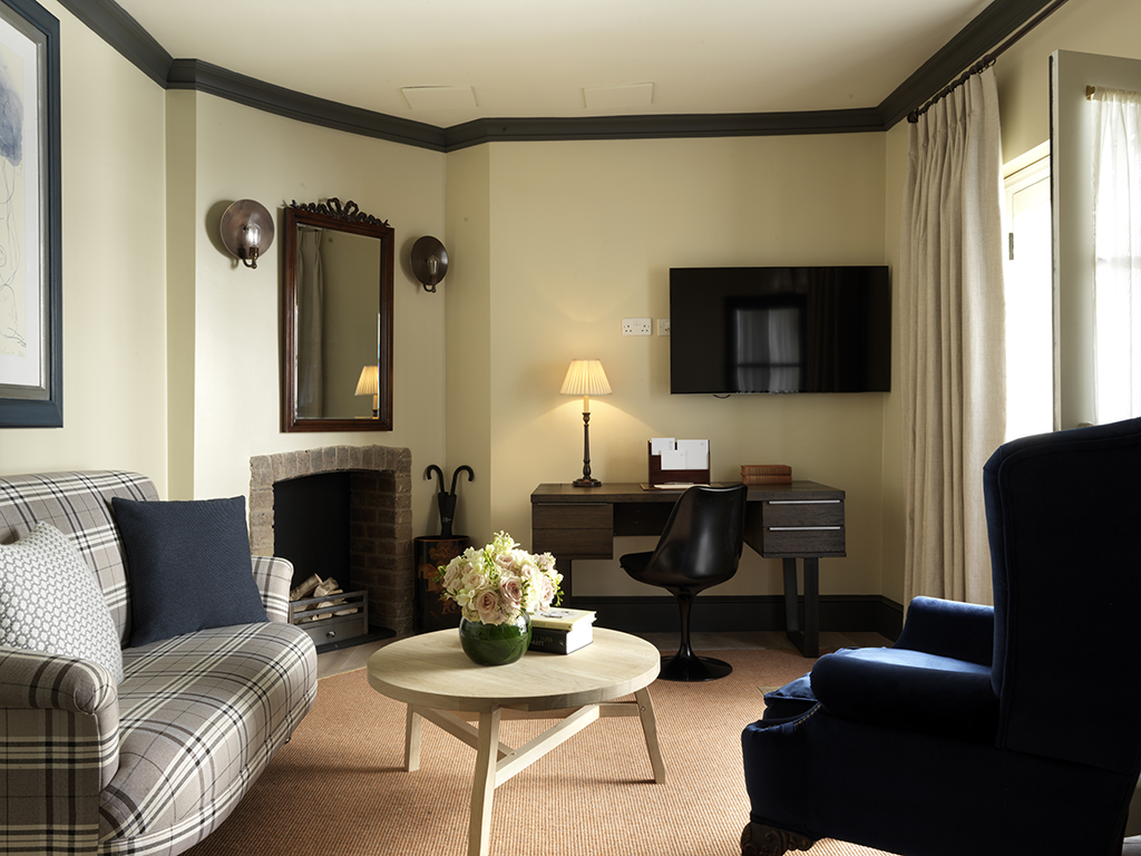 A cozy living room, perfect for Mother’s Day gatherings, features a plaid sofa, navy armchair, round coffee table with flowers, fireplace, wall mirror, desk with lamp and chair, wall-mounted TV, and beige curtains covering a window.