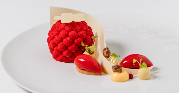 A sophisticated dessert inspired by the Chelsea Flower Show, featuring a red, textured sphere resembling raspberries, served with red and creamy dollops, garnishes, caramelized nuts, and a curved white chocolate decoration on a white plate.