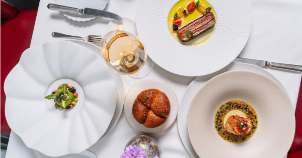 Three elegantly plated gourmet dishes, a round bread roll, and a glass of white wine arranged on a white tablecloth with cutlery, viewed from above—perfect for a sophisticated Chelsea Flower Show luncheon.
