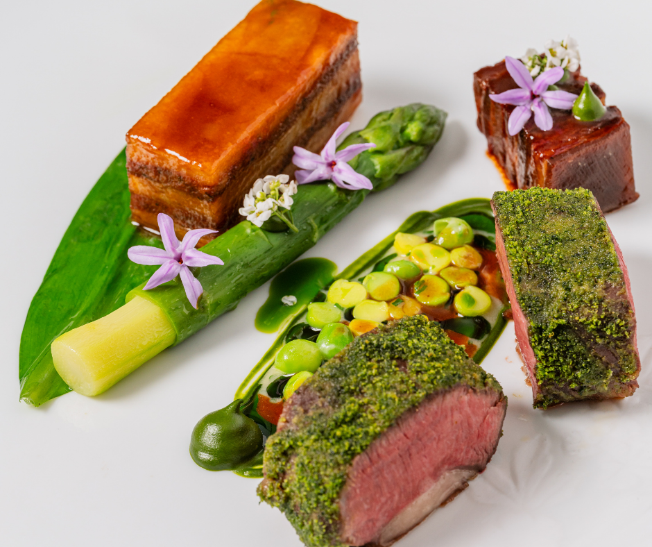 A gourmet dish from The Game Bird menus featuring seared meat slices, glazed pork belly, green vegetable puree, asparagus, and garnishes of edible flowers and leaves, artfully arranged on a white plate.