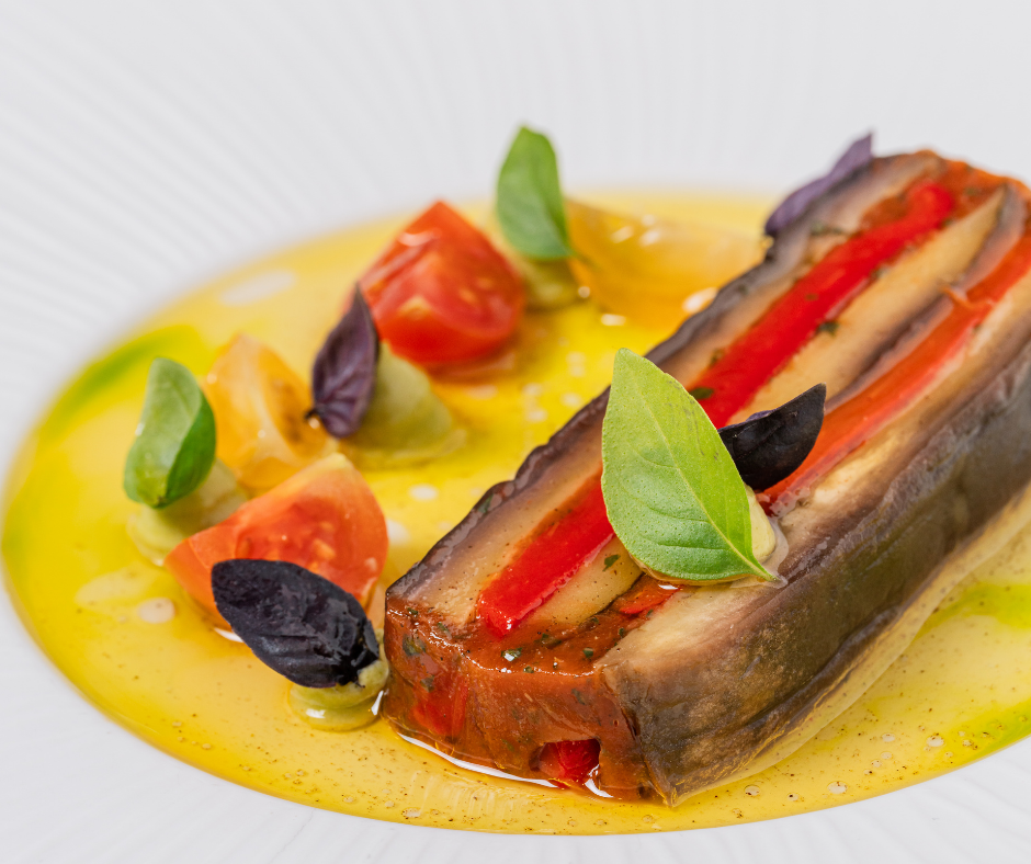 A plated dish from The Game Bird menus featuring a layered vegetable terrine with eggplant, zucchini, and red pepper, garnished with basil leaves and cherry tomato halves, surrounded by a yellow sauce.