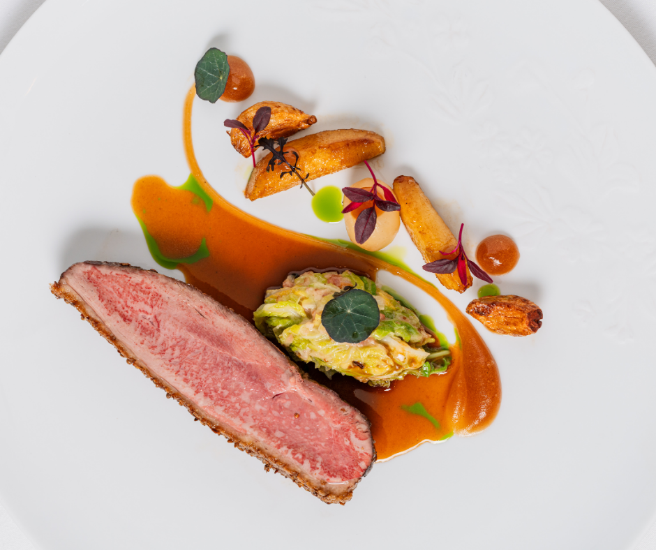 A gourmet dish inspired by The Game Bird menus, featuring a seared slice of medium-rare meat, roasted potatoes, vegetables topped with a green leaf, and drizzles of brown sauce and herb oil, artfully arranged on a white plate.