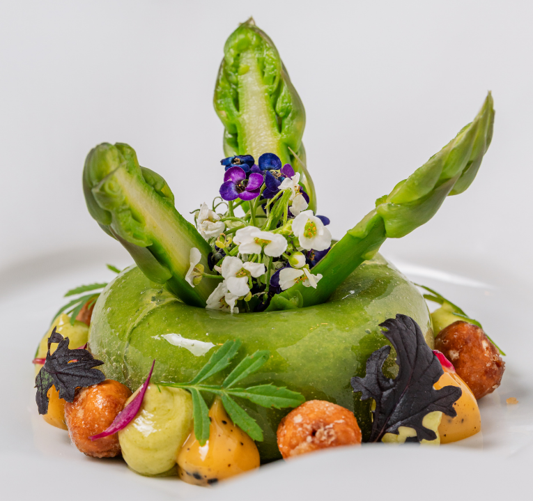 A gourmet dish from The Game Bird menus featuring three upright asparagus spears set in a green mousse ring, garnished with edible flowers, small vegetables, leafy greens, and caramelized nuts on a white plate.