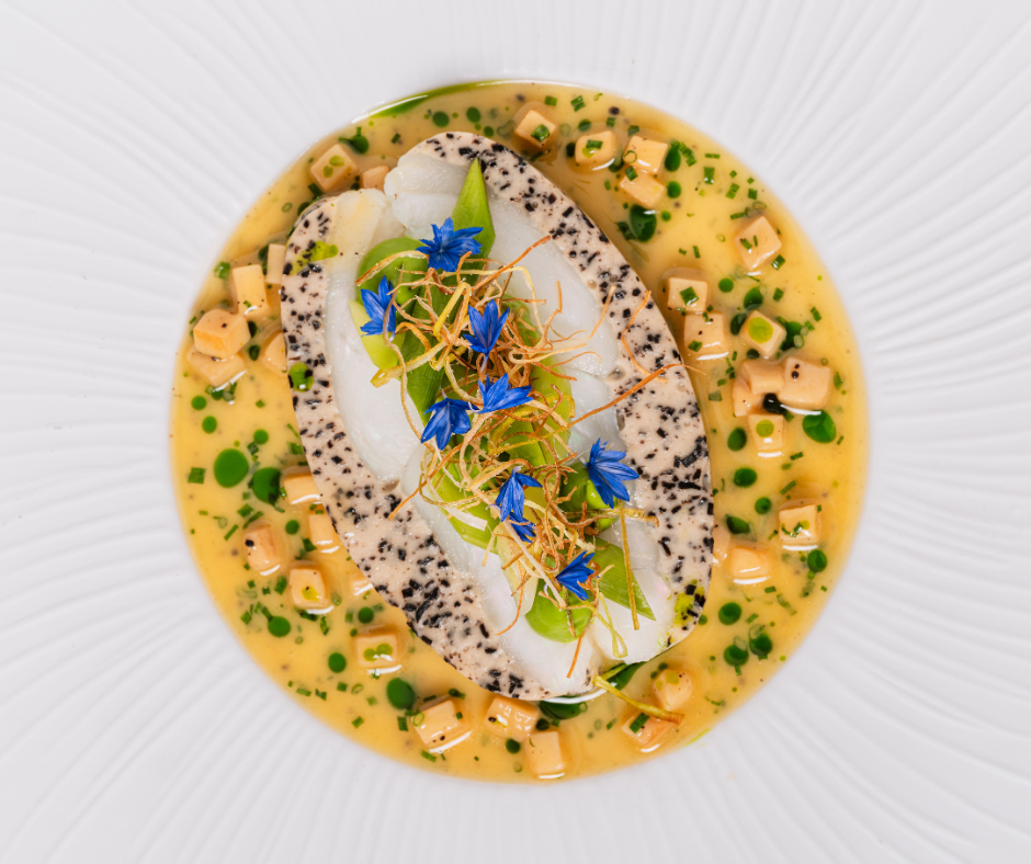 A gourmet dish from Michael Caines at The Stafford features two slices of white fish with herbs and black specks, garnished with blue edible flowers and greens, served in a creamy yellow sauce with diced vegetables.