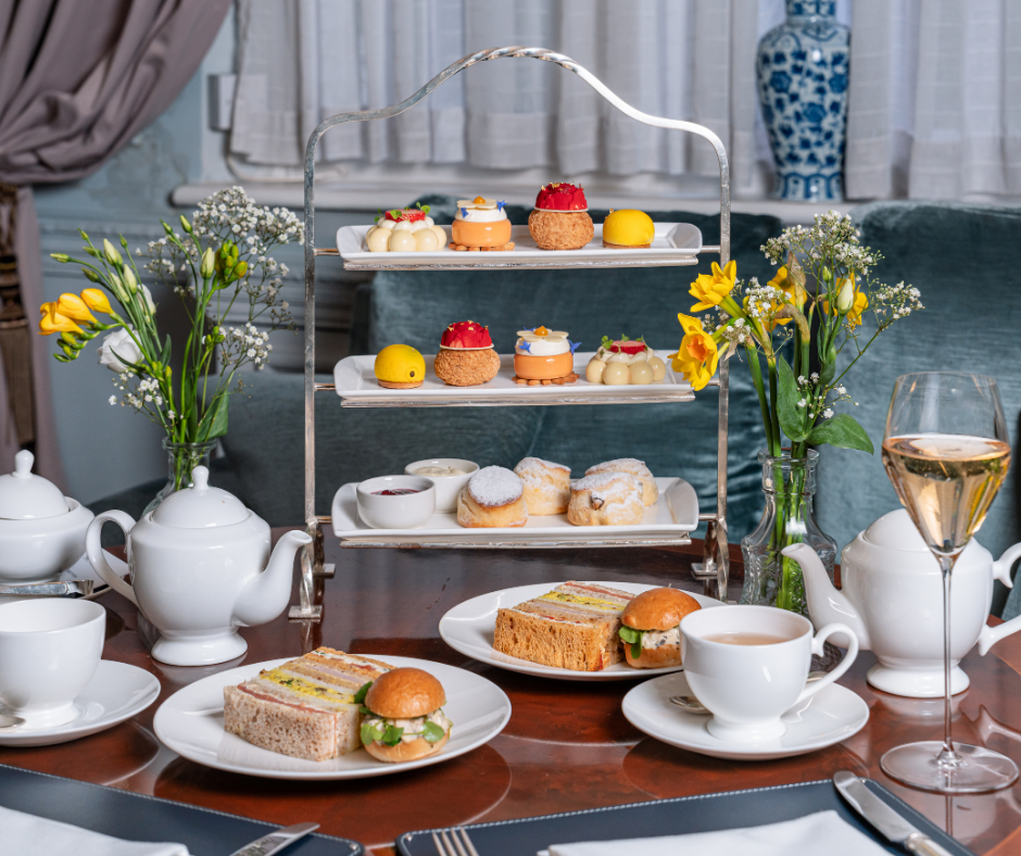 An elegant afternoon tea setup at Michael Caines at The Stafford with tiered trays of assorted pastries and desserts, plates of sandwiches and sliders, teapots, cups, a glass of champagne, and vases of yellow and white flowers on a wooden table.