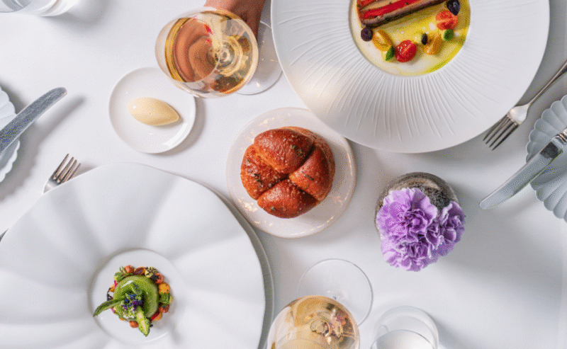 A top-down view of a fine dining table at Michael Caines at The Stafford, with two people toasting white wine, elegant plated dishes, a bread roll, butter, and a purple flower centerpiece on a white tablecloth.