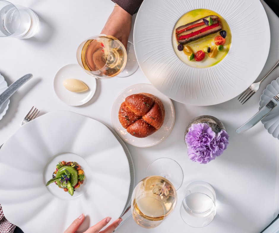 A top-down view of a fine dining table at Michael Caines at The Stafford, with two people toasting white wine, elegant plated dishes, a bread roll, butter, and a purple flower centerpiece on a white tablecloth.