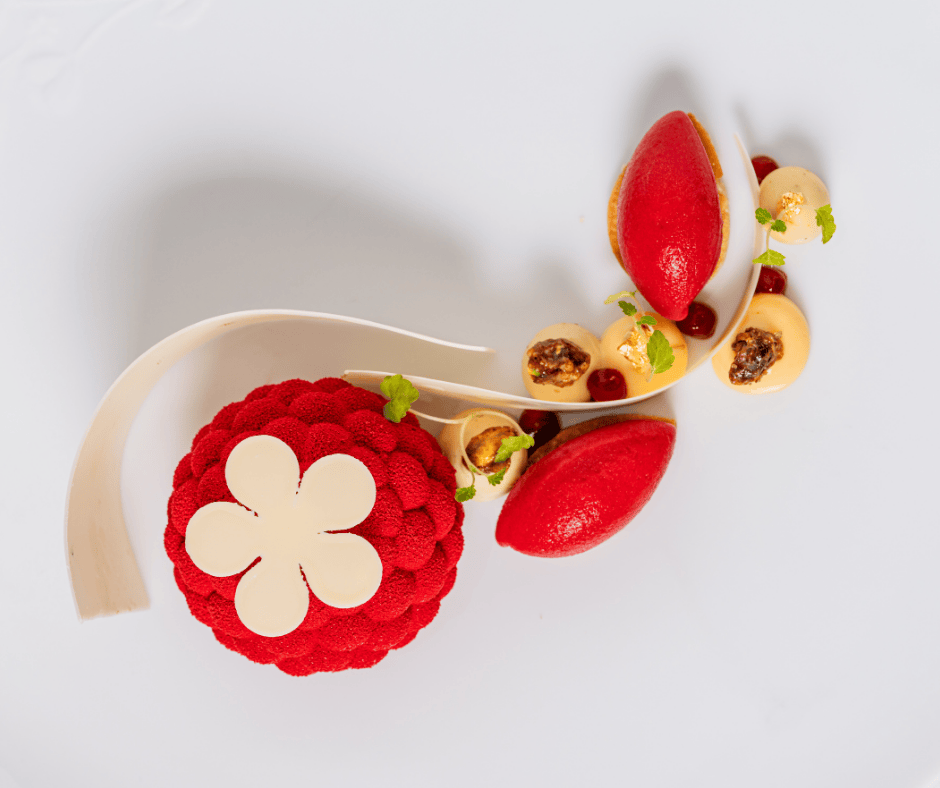 A gourmet dessert at Michael Caines at The Stafford features a red, textured dome topped with a white chocolate flower, two red quenelles, creamy dollops, caramelized nuts, small green herbs, and a curved white chocolate decoration on a white plate.