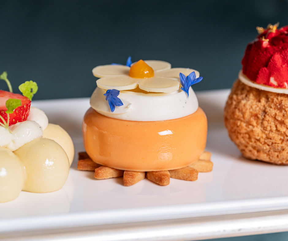 A close-up of three elegant pastries on a white plate, perfect for afternoon tea. The center pastry is orange with white cream, almond petals, blue flower petals, and a yellow center, resting on a crisp base.
