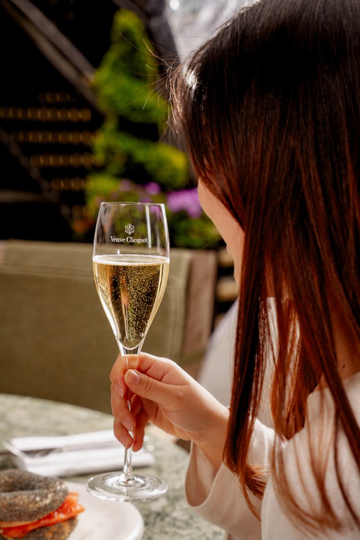 A woman with long hair holds a glass of Veuve Clicquot champagne at a table in the Enemigo Tequila Courtyard, with food partially visible in the foreground and blurred greenery in the background.