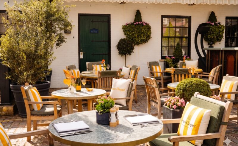 An outdoor café patio, the Enemigo Tequila Courtyard, features round tables, wooden chairs with yellow-striped cushions, potted plants, flowers, and greenery on a cobblestone floor before a white building with black-trimmed windows and a green door.