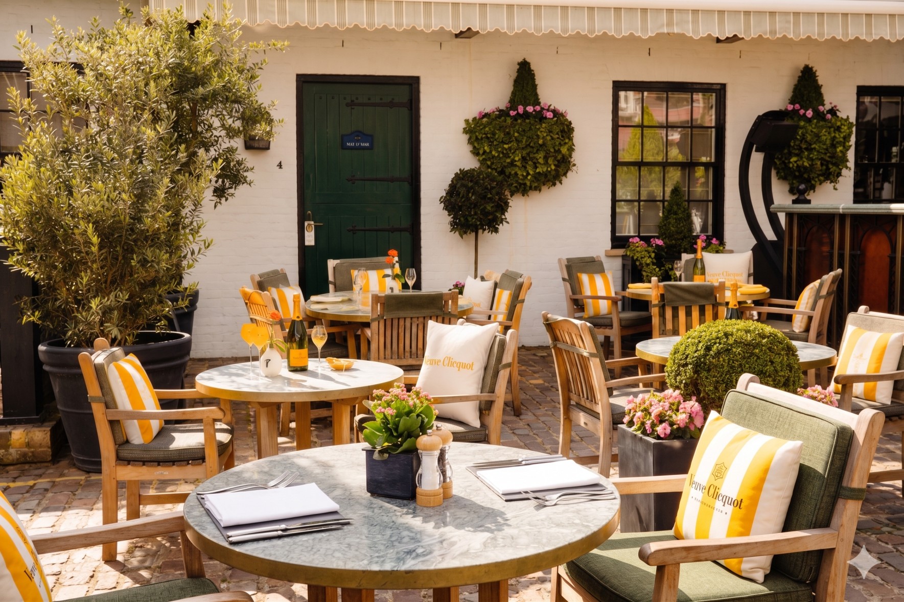 An outdoor café patio, the Enemigo Tequila Courtyard, features round tables, wooden chairs with yellow-striped cushions, potted plants, flowers, and greenery on a cobblestone floor before a white building with black-trimmed windows and a green door.