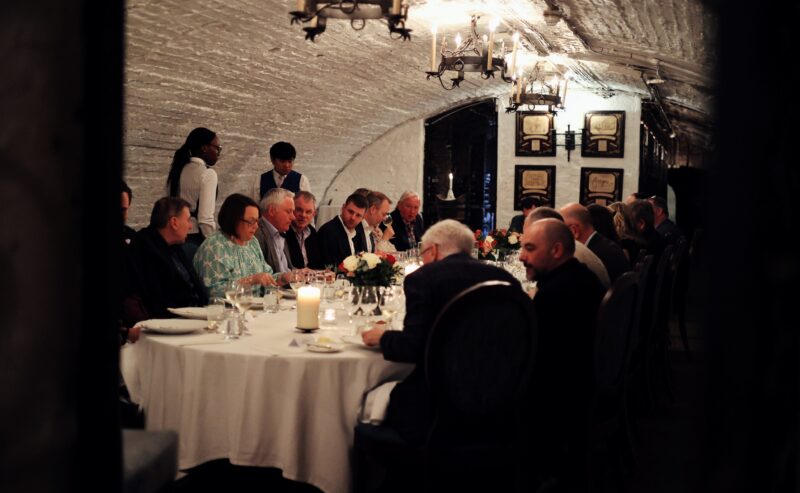 A group of people sit around a long, elegantly set dining table in a dimly lit, arched brick room. Candles and floral centerpieces decorate the table, creating a formal and intimate dinner atmosphere.