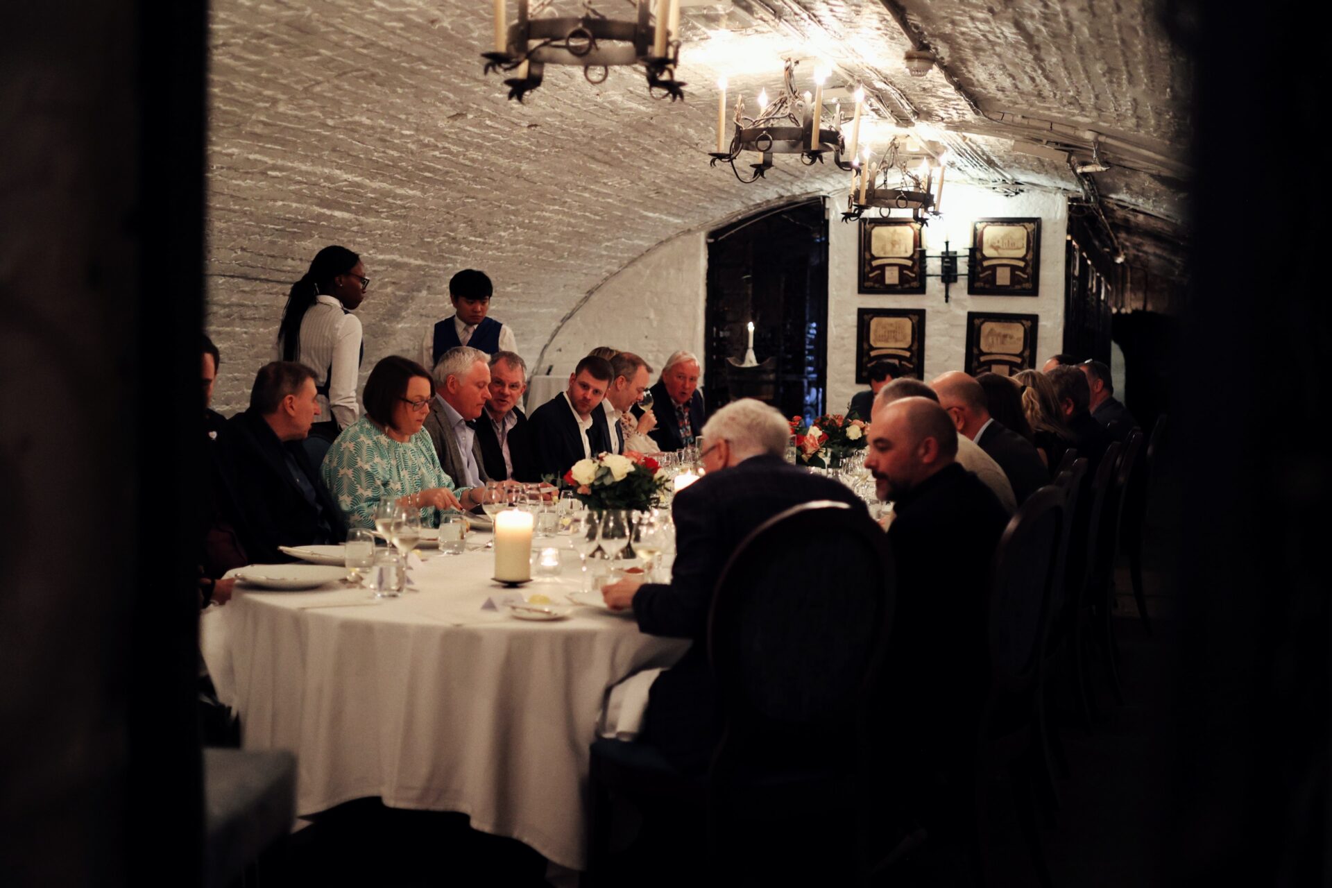 A group of people sit around a long, elegantly set dining table in a dimly lit, arched brick room. Candles and floral centerpieces decorate the table, creating a formal and intimate dinner atmosphere.