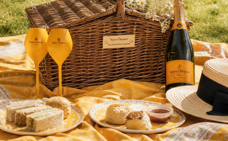 A wicker picnic basket with white flowers, two Veuve Clicquot champagne glasses, a bottle of Veuve Clicquot, and an array of treats are set on a yellow patterned blanket outdoors—perfect for a Chelsea Flower Show-inspired afternoon.